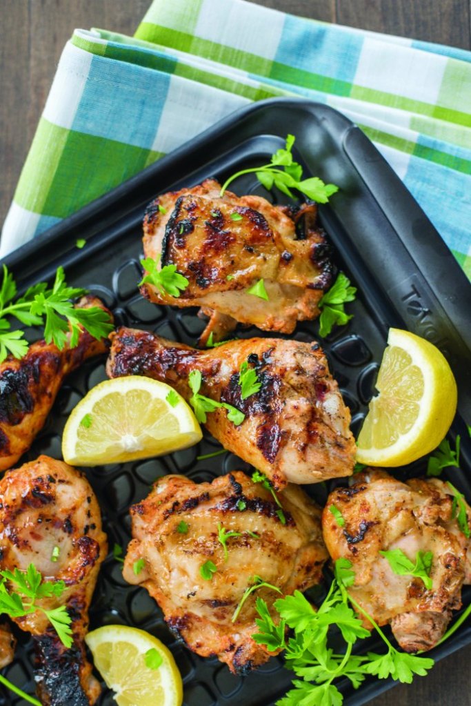 Grilled chicken thighs garnished with fresh parsley and lemon slices on a black tray with a green and blue checkered napkin in the background.