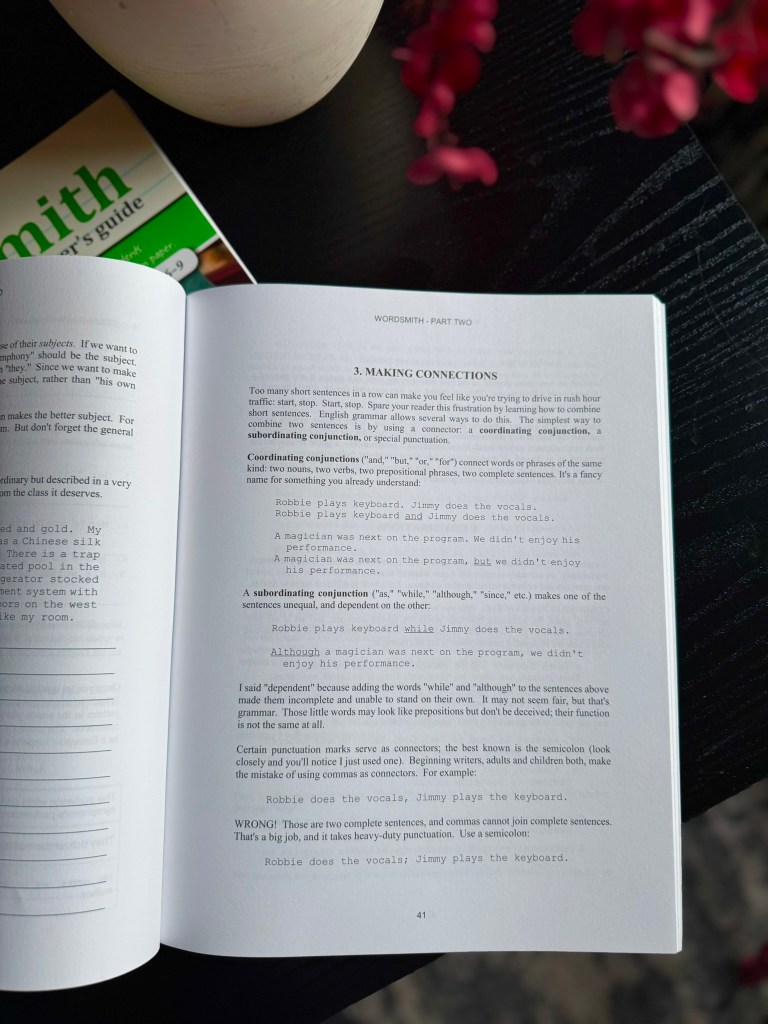 An open book titled 'Wordsmith - Part Two', displaying a page about making connections in writing, with examples of coordinating and subordinating conjunctions, on a dark wooden table next to a decorative vase and a green book.