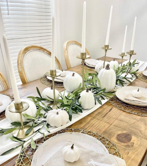 A beautifully set fall table featuring soft white pumpkins, elegant gold candlesticks with white candles, and green foliage arranged along the center. The table is adorned with neutral-toned plates, creating a warm and inviting atmosphere.