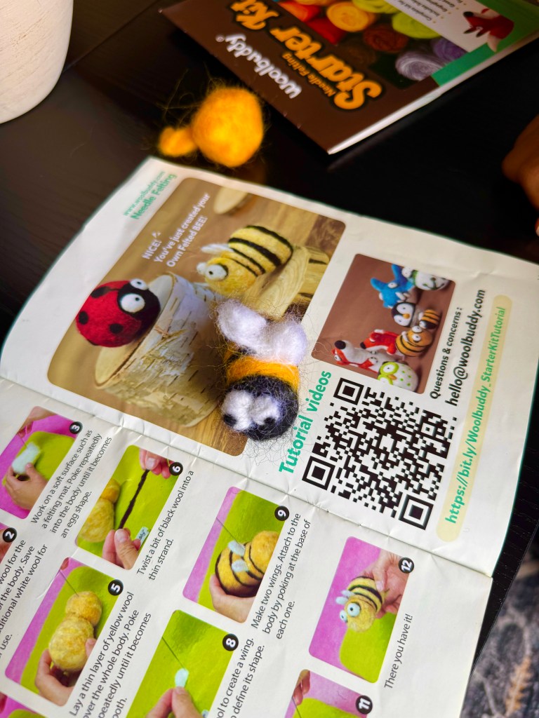 A close-up of a needle-felted bee figure resting on a colorful instructional booklet from the Woolbuddy Needle Felting Starter Kit, showcasing step-by-step tutorials for crafting projects.