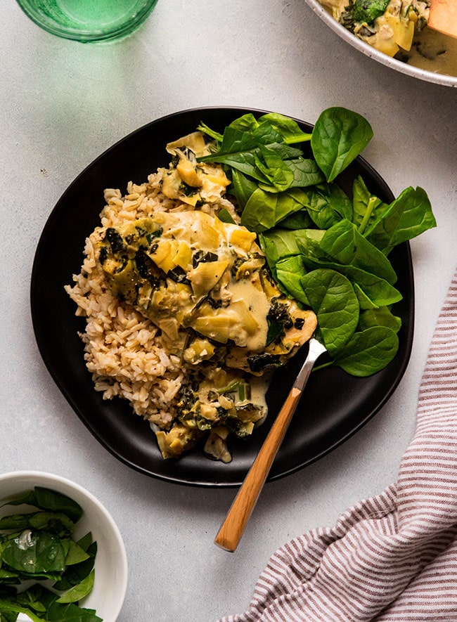 A serving of creamy garlic chicken with artichokes, served over brown rice, accompanied by fresh spinach on a black plate.