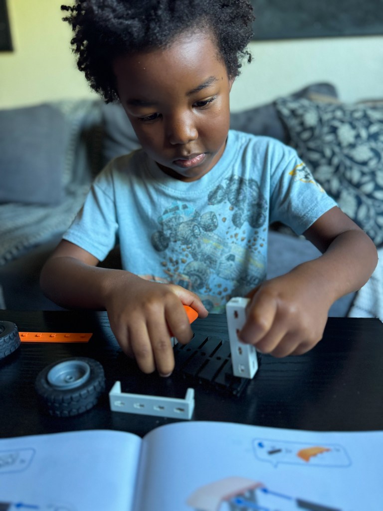 A young child with curly hair focused on assembling a toy, using building blocks, wheels, and a manual on a table.