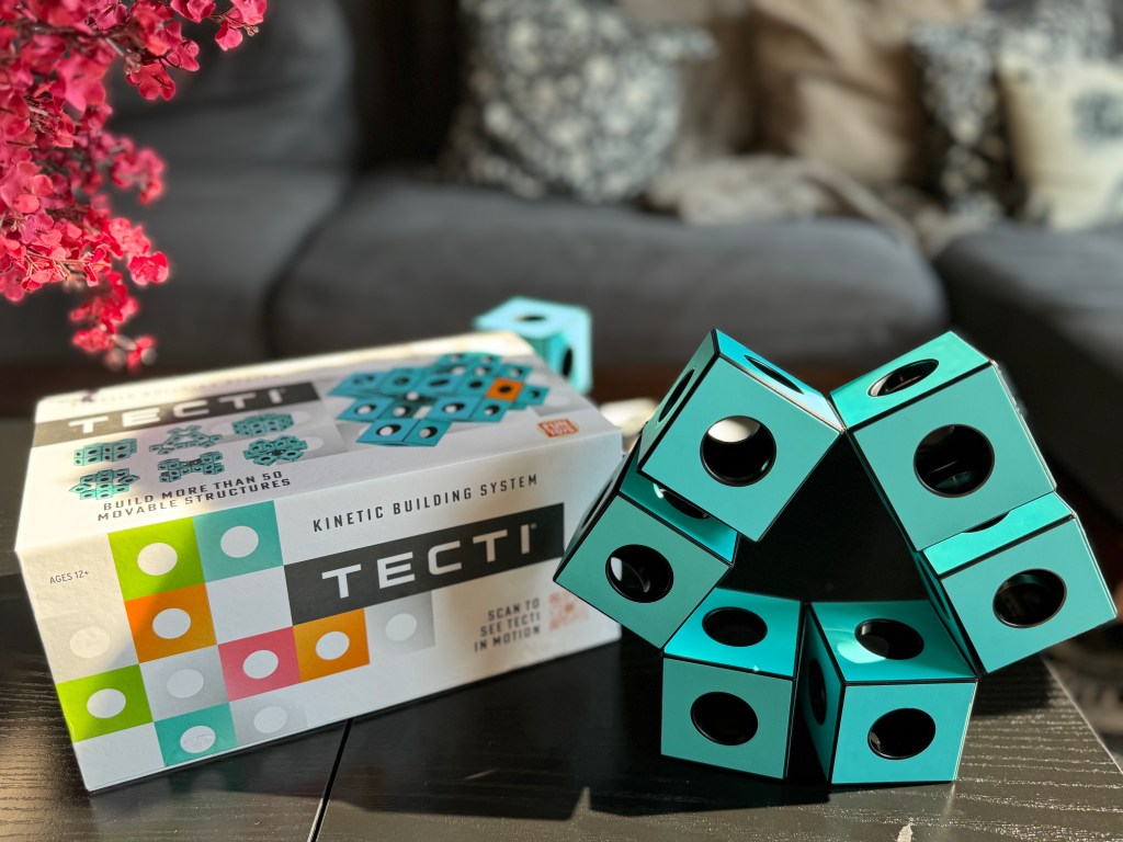 A close-up image of the Tecti kinetic building system box featuring turquoise cubes arranged in a dynamic structure on a dark table with pink flowers in the background.
