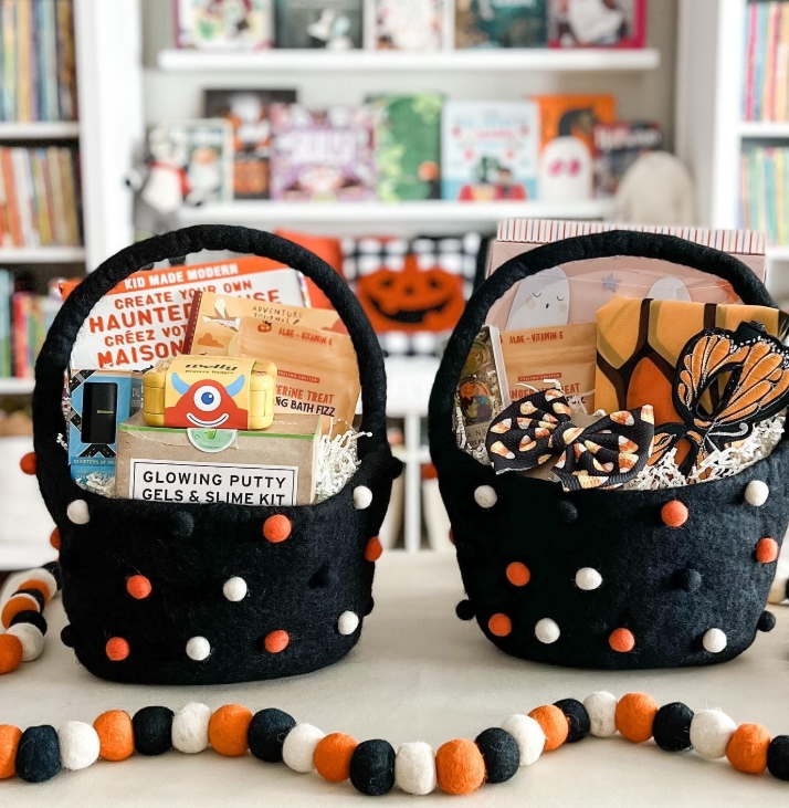 Two black felt baskets decorated with colorful pom-poms are filled with Halloween-themed treats, including glowing putty, a slime kit, and craft items. A playful garland of felt balls is displayed in the foreground.