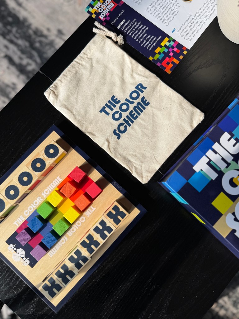 An overhead view of 'The Color Scheme' board game, featuring a canvas storage pouch, colorful wooden blocks, and game components displayed on a black table.