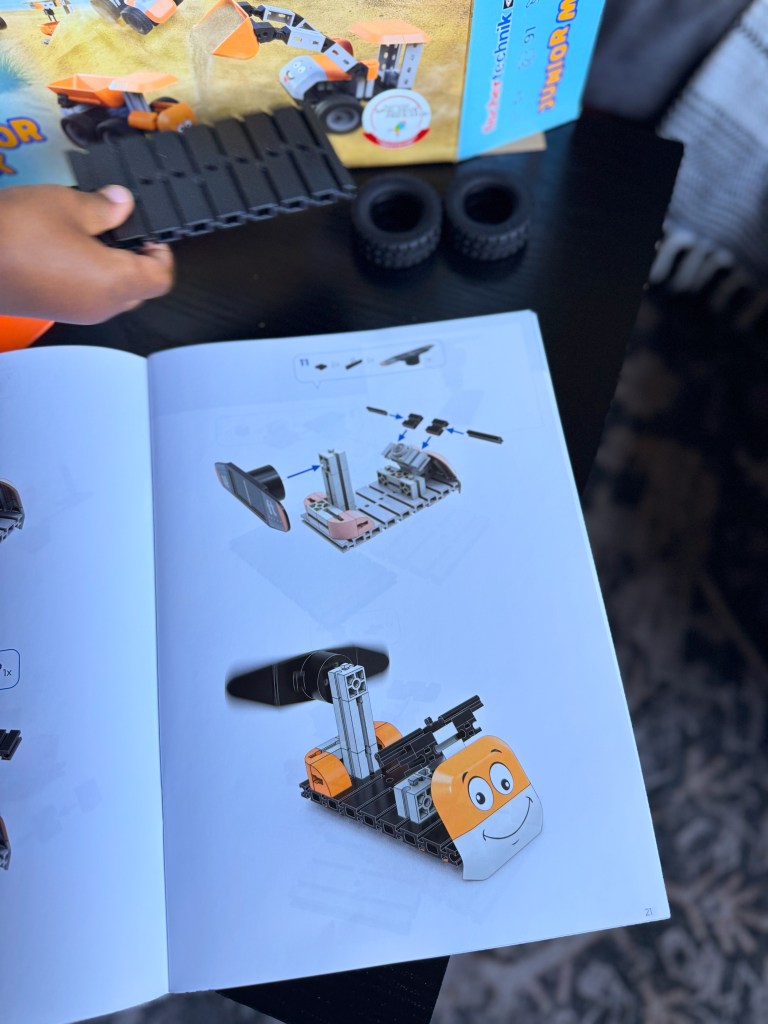 A close-up image showing a child’s hand assembling a toy construction vehicle using black tracks, with a colorful instruction manual open to a page illustrating the vehicle's assembly, and two black wheels visible beside it.