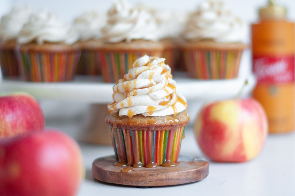 A close-up of a beautifully decorated cupcake topped with swirls of frosting and drizzled with caramel, surrounded by fresh apples and colorful cupcake wrappers.