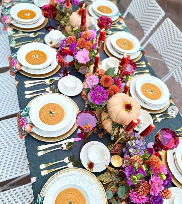 An elegantly styled Thanksgiving tablescape featuring white dinnerware, rich autumn floral arrangements, small pumpkins, and vibrant red candles. The table is beautifully set with gold flatware and colorful accents, ready for a festive gathering.