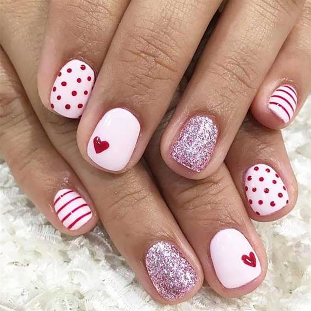 Close-up of manicured nails featuring various pink and white designs, including polka dots, stripes, and a heart, with one nail showcasing silver glitter.