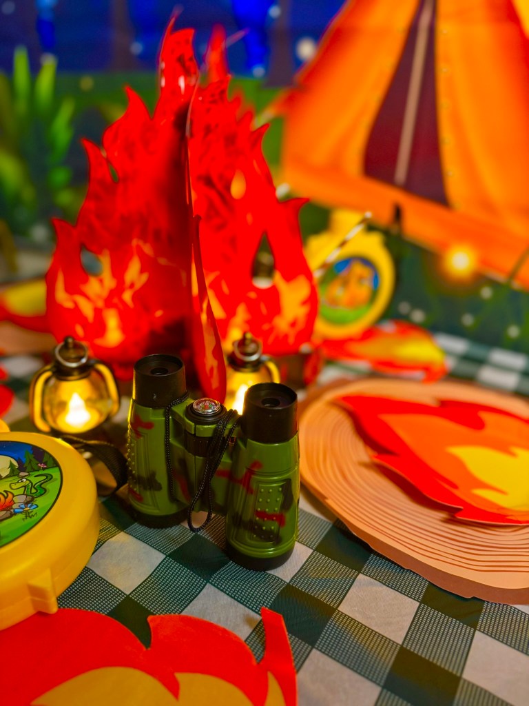 Kids camping party decorations with green binoculars, yellow canteens, and glowing lanterns on a gingham tablecloth.
