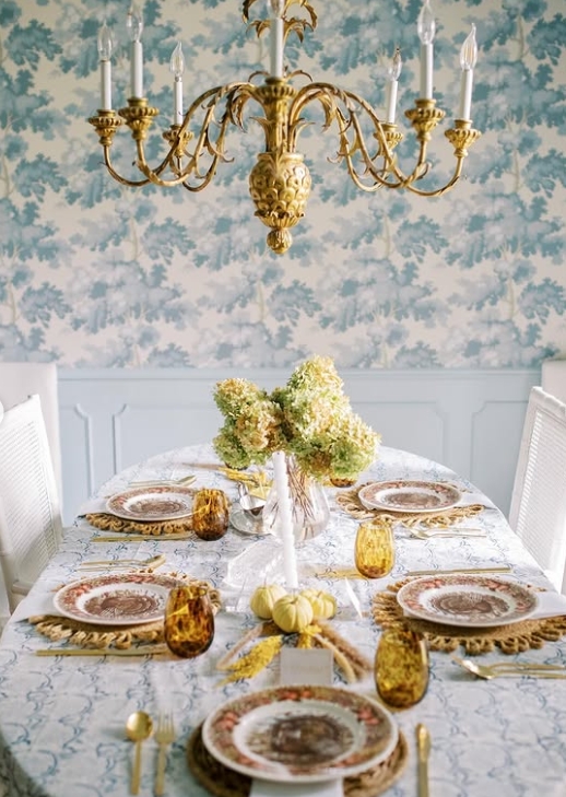 An elegant Thanksgiving tablescape featuring a round table with floral dinnerware, amber glassware, and a centerpiece of fresh hydrangeas. A chandelier hangs above and a light blue wallpaper creates a warm ambiance.