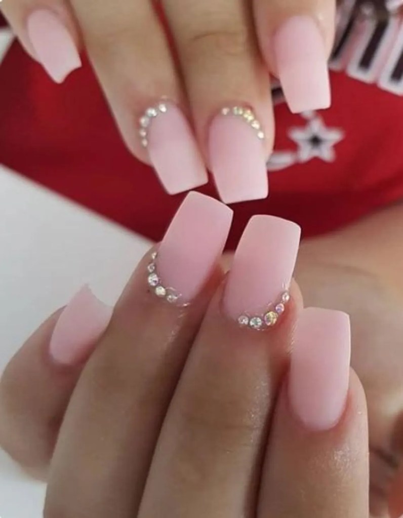 Close-up of hands displaying long, square-shaped nails with a matte pink finish and embellished with small rhinestones.