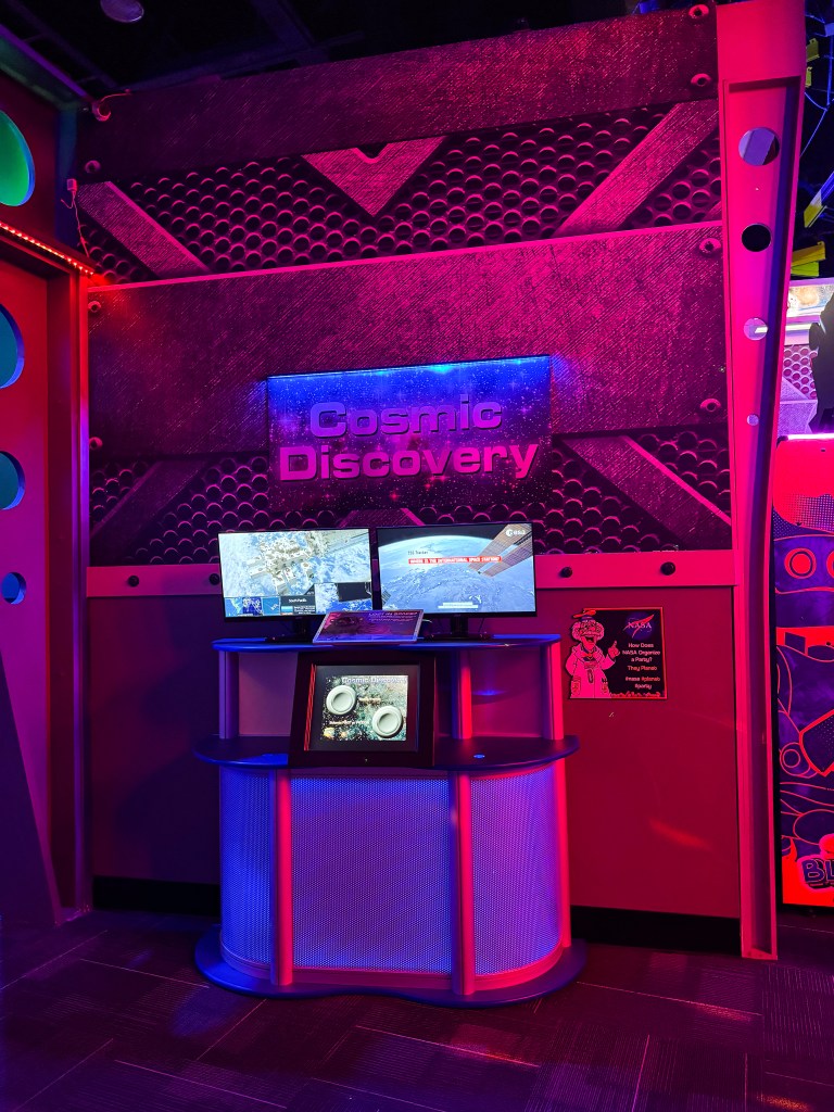 An interactive display titled 'Cosmic Discovery' featuring two screens showing space-related content, set against a vibrant pink and metallic backdrop.
