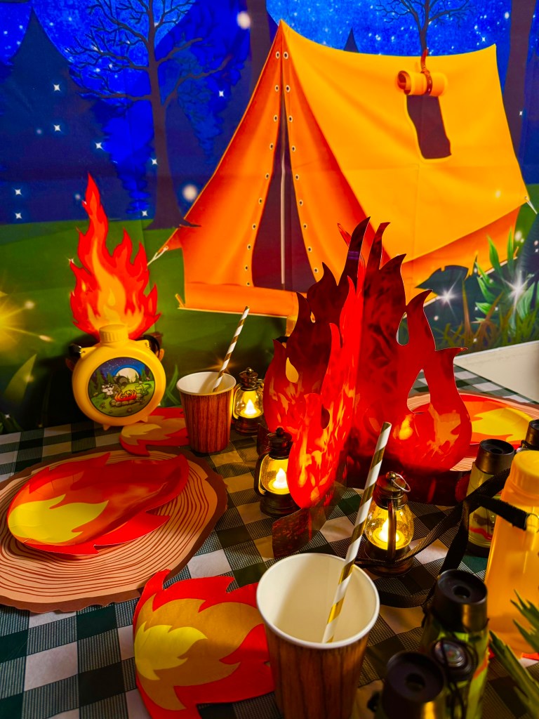 Camping birthday party campfire centerpiece with red flames, lanterns, and woodland table decor.
