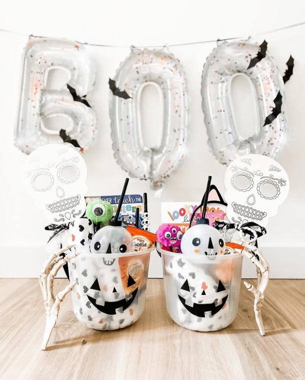 Two Halloween-themed baskets filled with treats and toys, featuring smiling skulls and decorative items, displayed against a backdrop of large silver 'BOO' balloons.