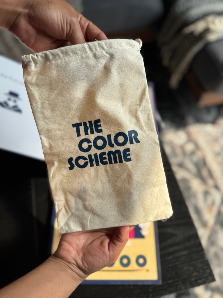 A person holding a canvas pouch labeled 'THE COLOR SCHEME,' with a game board partially visible in the background.