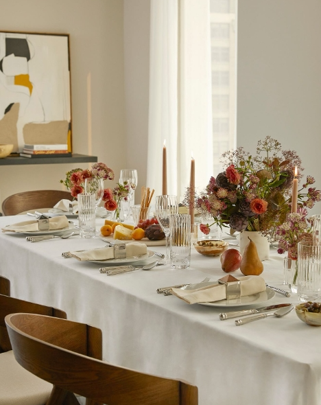 A beautifully set dining table adorned with a white tablecloth, elegant glassware, and a floral centerpiece featuring autumn-colored blooms. The setting includes candles, assorted fruits, and decorative elements, creating a warm and inviting atmosphere for a fall gathering.