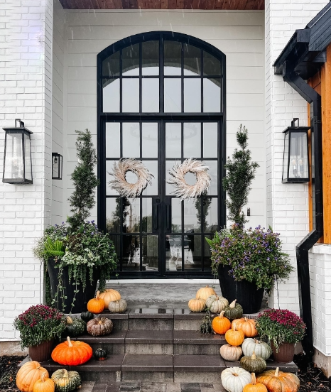 A stylish porch featuring double black doors adorned with natural wreaths. The entryway is decorated with an array of colorful pumpkins and lush greenery in pots, creating a cozy autumn atmosphere.