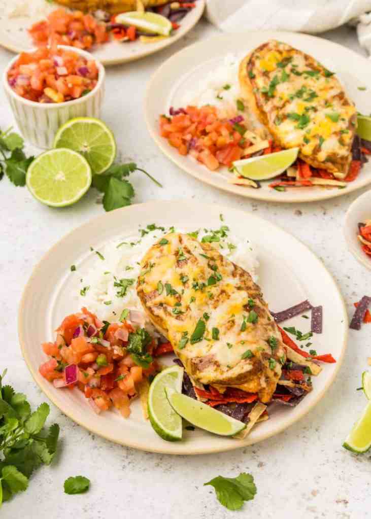 A plate of baked chicken with melted cheese, served with white rice, colorful pico de gallo, lime wedges, and fresh cilantro garnish.