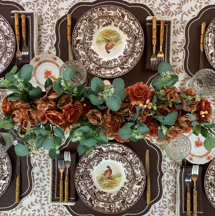 Elegant Thanksgiving tablescape featuring rich floral arrangements with warm red tones, vintage-inspired dinnerware, and natural textures. The setting includes layered plates, glassware, and utensils, creating an inviting atmosphere.