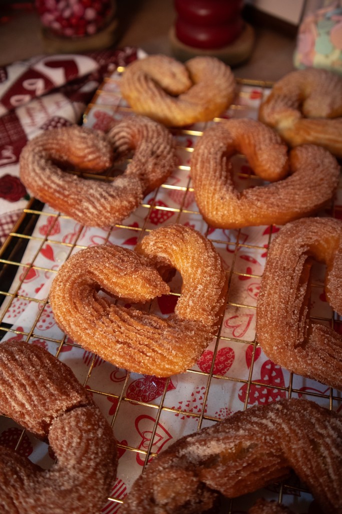 A tray of heart-shaped, sugar-coated pastries cooling on a wire rack, with a decorative cloth underneath.