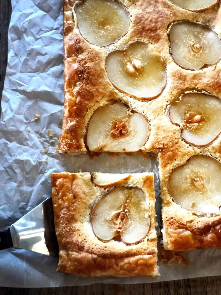 A freshly baked pear tart with golden-brown pastry and thinly sliced pears on top, partially sliced and served on parchment paper.