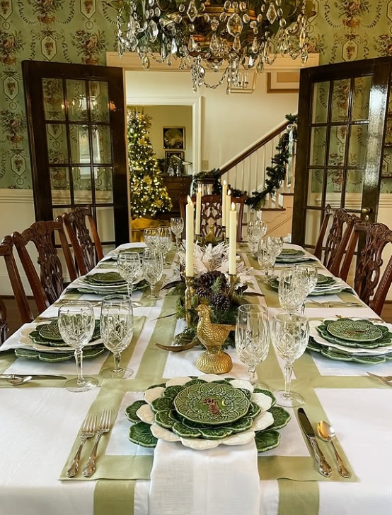 A beautifully styled Thanksgiving table featuring green floral dinnerware, crystal glassware, and gold accents, with a centerpiece of pinecones and candles. The setting is elegant, complemented by a chandelier and festive decorations in the background.
