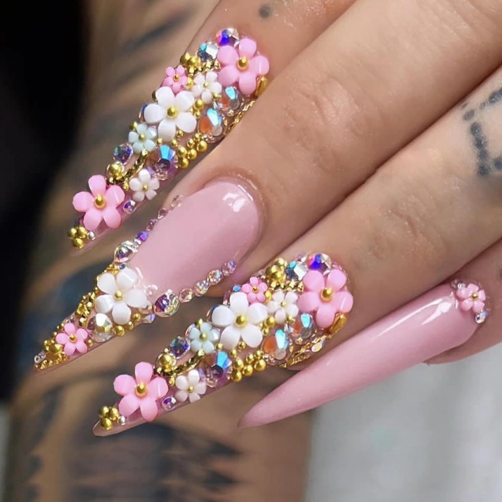 Close-up of elegantly manicured long nails adorned with pink and white flowers, gold beads, and colorful rhinestones, showcasing a decorative and stylish nail art design.