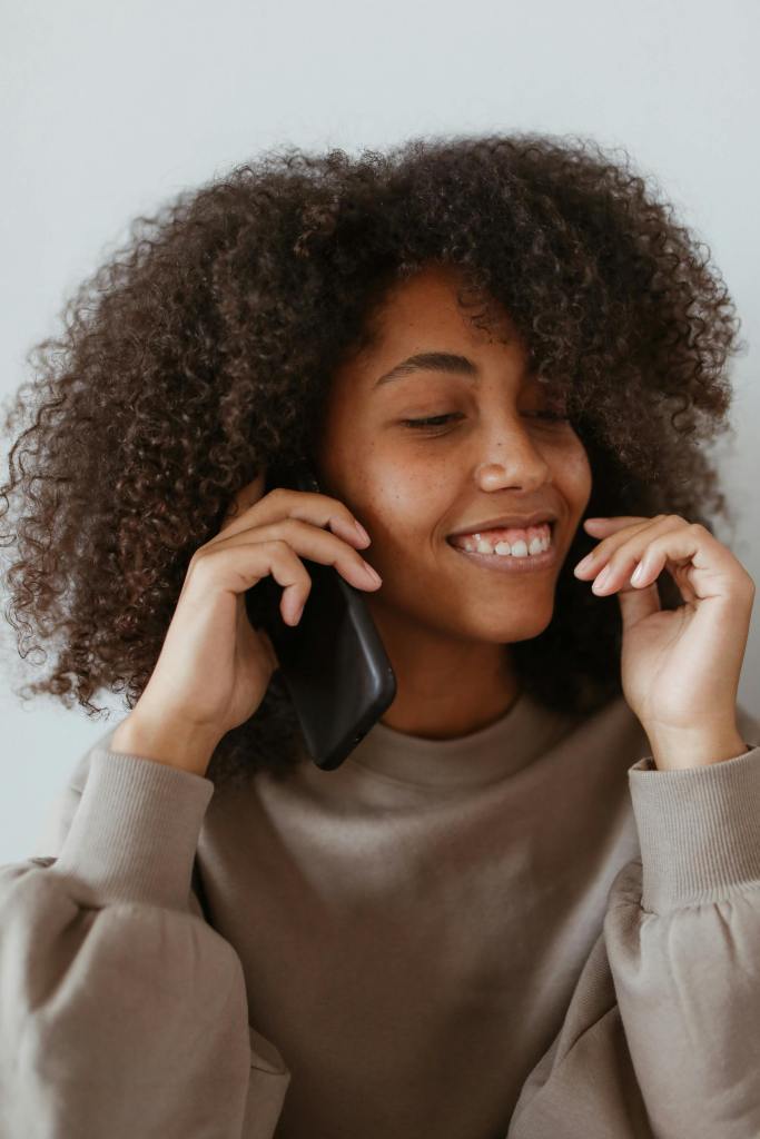 Smiling woman with curly hair talking on the phone, wearing a beige sweater.
