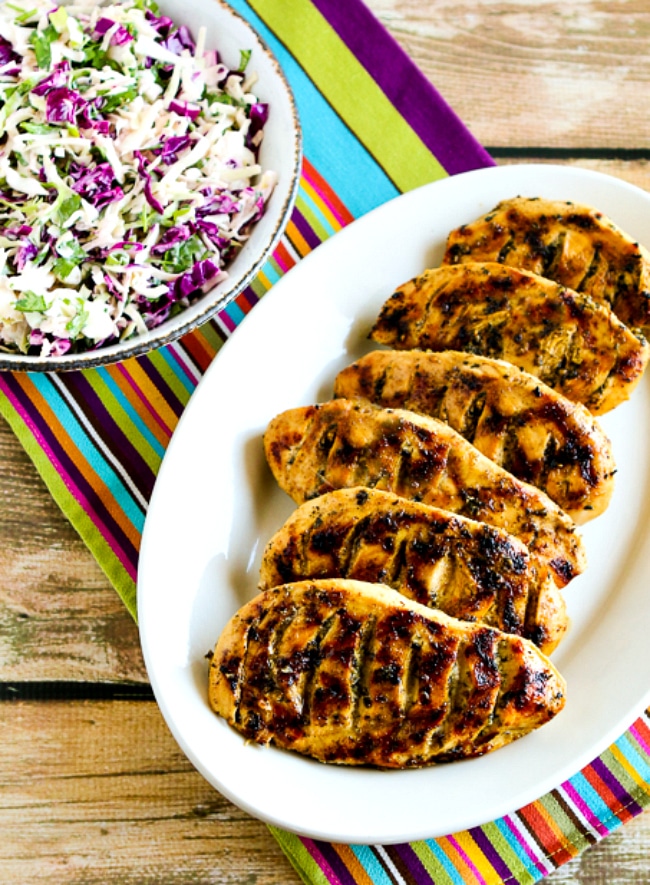 Grilled chicken breasts served on a white platter next to a bowl of colorful chopped coleslaw, set on a striped tablecloth.
