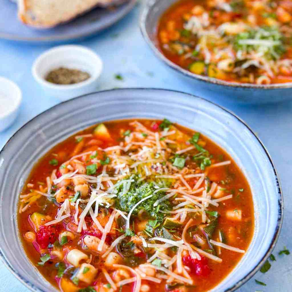 A bowl of hearty vegetable soup topped with shredded cheese and fresh herbs, served alongside a slice of bread.