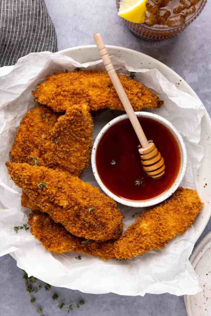 Crispy baked chicken tenders arranged on a plate with a small bowl of sweet sauce and a honey dipper, garnished with fresh herbs.