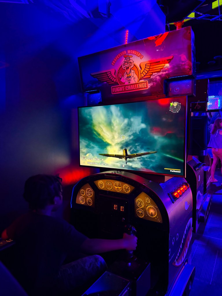 A child playing an arcade flight simulator game, sitting at the controls of a colorful cockpit with a large screen displaying a flying scene and game objectives, surrounded by ambient colorful lighting.
