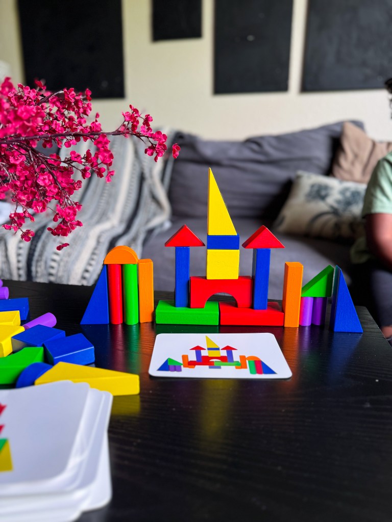 A colorful arrangement of building blocks on a black table, featuring various geometric shapes forming a castle-like structure, with a card showing the design beside it. A decorative plant with pink flowers is in the foreground, and a person is blurred in the background.
