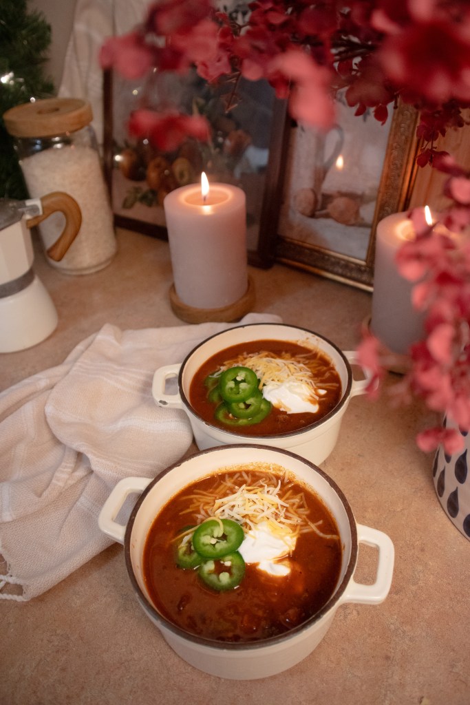 Two bowls of vegan mushroom chili topped with sliced jalapeños, shredded cheese, and sour cream, placed on a countertop with candles and decorative elements in the background.