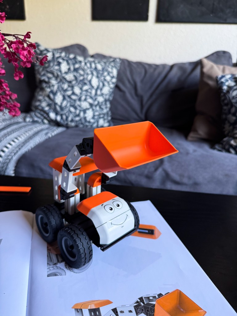 A colorful toy construction vehicle with a smiling face, featuring an orange scoop and black wheels, positioned on an open instruction manual on a black table, with decorative pillows and a pink flower pot in the background.