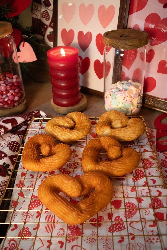 A decorative display of heart-shaped pastries on a golden wire rack, surrounded by festive Valentine's Day decorations, including a red candle and jars of colorful candies.