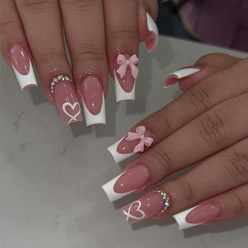Close-up of manicured hands featuring long nails with a pink base and white tips. Some nails have pink bows and heart designs, along with rhinestone embellishments.