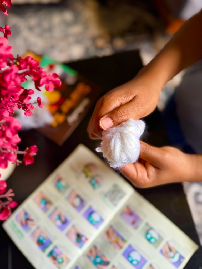 A child's hand holding a clump of white wool while following instructions from a colorful needle felting kit manual, with a background of pink flowers.