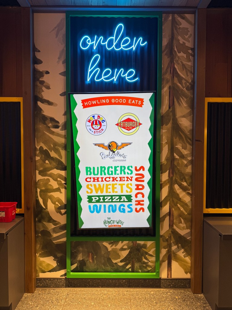 Signboard displaying food options at Great Wolf Lodge including burgers, chicken, pizza, snacks, and sweets, with neon 'order here' sign above.