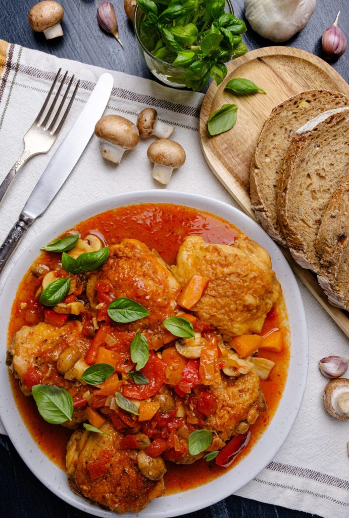 A delicious plate of chicken thighs cooked with colorful bell peppers, mushrooms, and fresh basil in a rich tomato sauce, served with slices of bread and garnished with herbs.