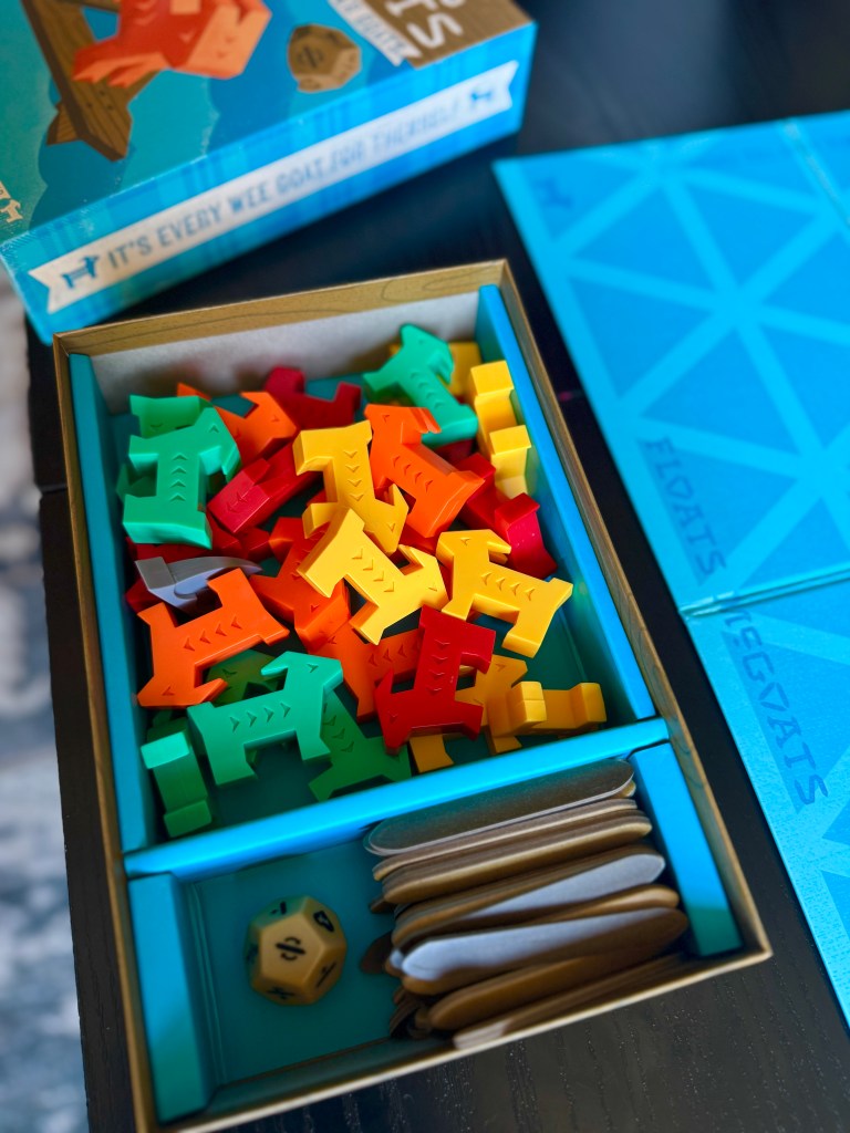A colorful assortment of game pieces in various animal shapes, including green, orange, yellow, and red, inside a game box. A blue game board is visible in the background with geometric patterns, alongside a set of scoring tokens and a die.