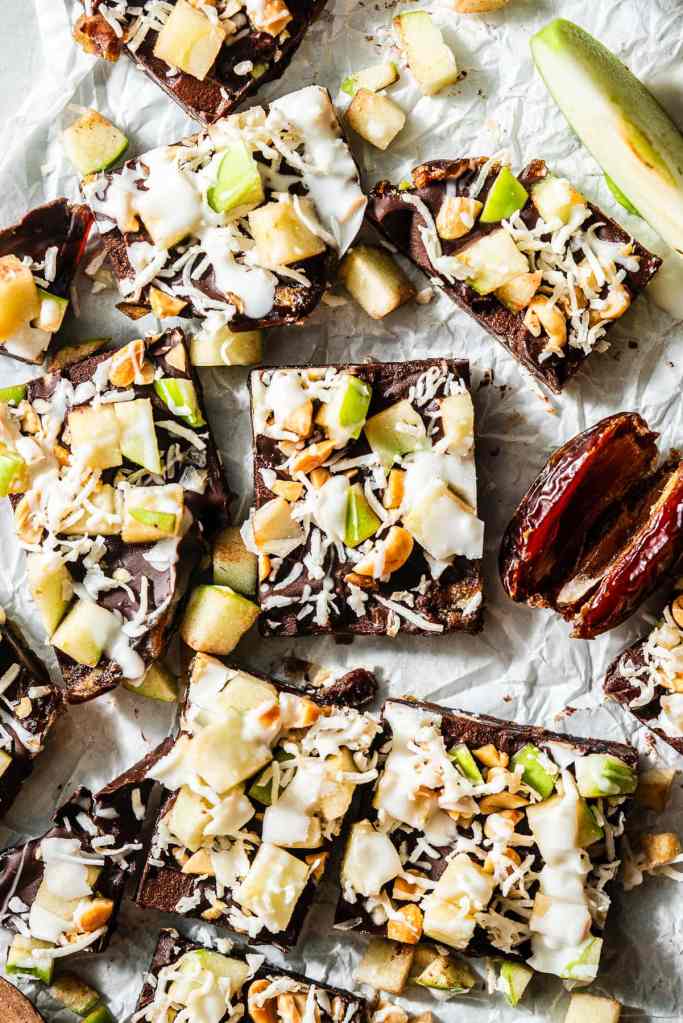 A close-up of chocolate date bark topped with diced apple, shredded coconut, and chopped nuts, arranged on crinkled parchment paper.