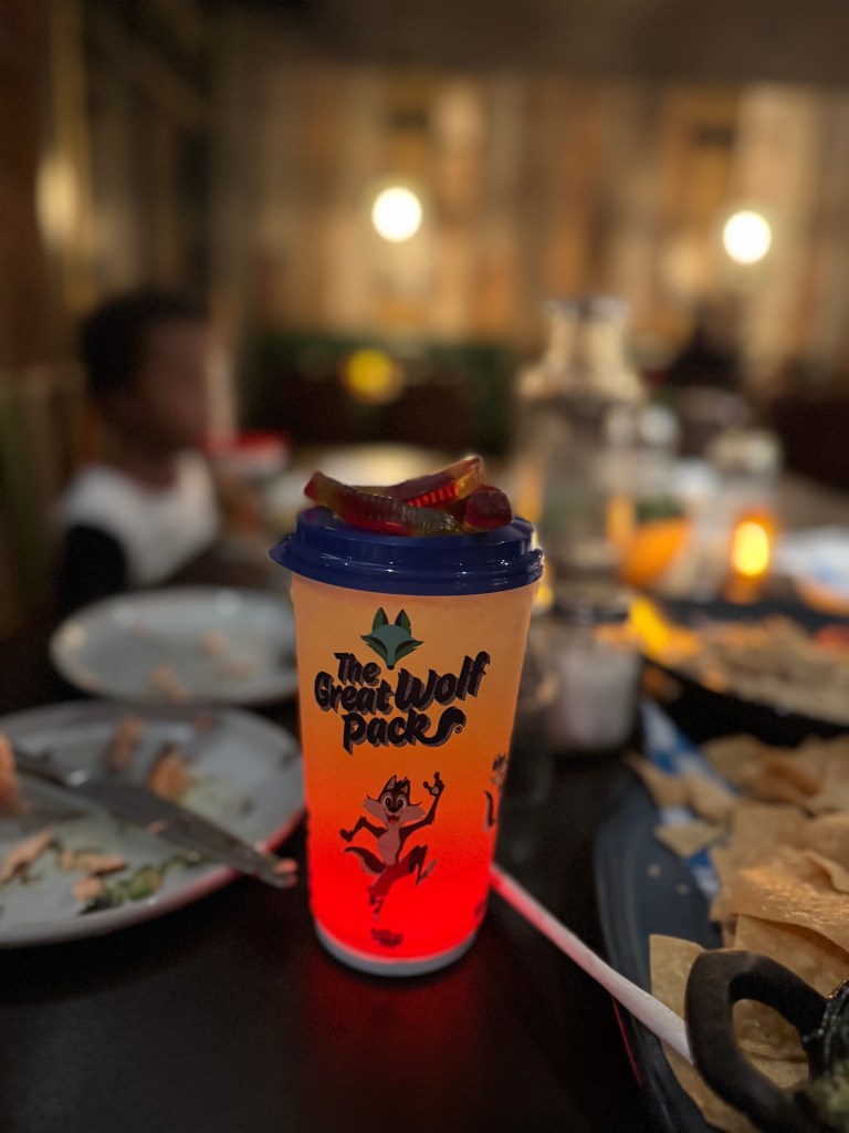 A colorful cup featuring 'The Great Wolf Pack' logo, filled with a gradient drink and topped with gummy candies, set against a dimly lit restaurant table with plates of food.