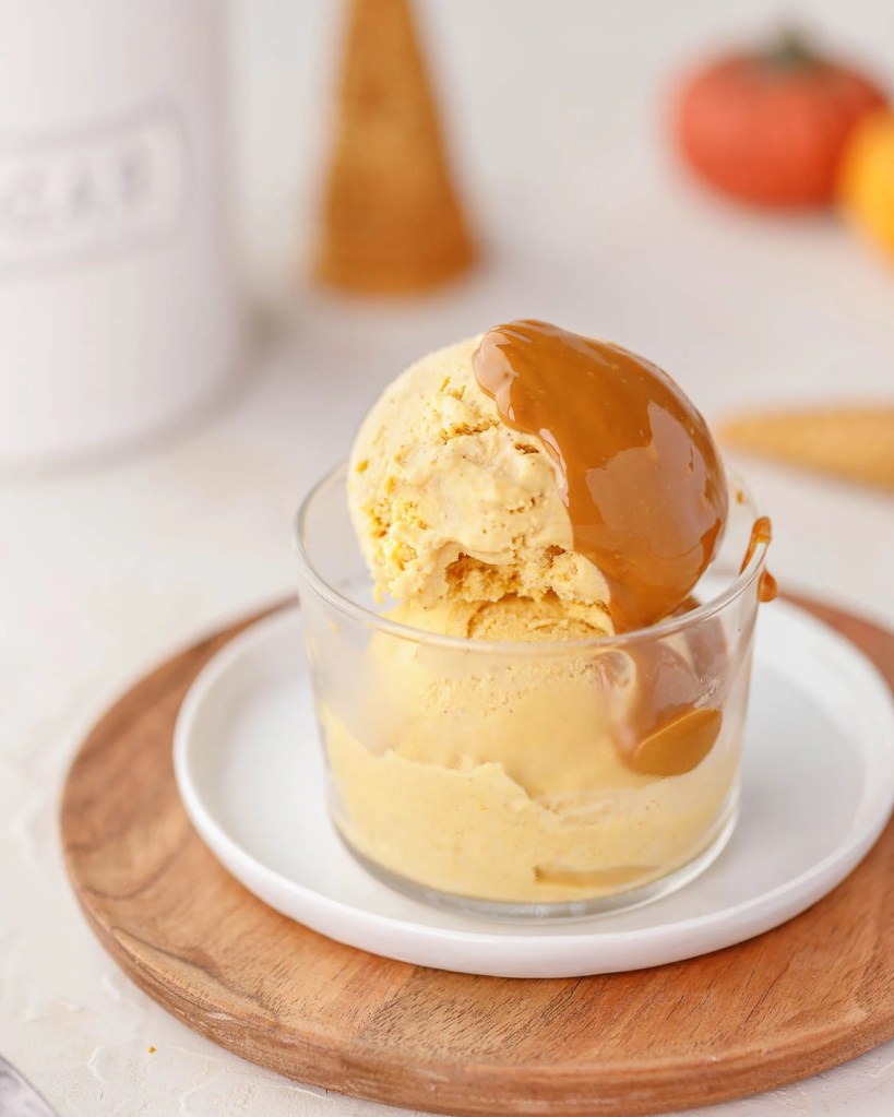 A glass bowl filled with creamy pumpkin ice cream drizzled with caramel sauce, placed on a wooden tray.