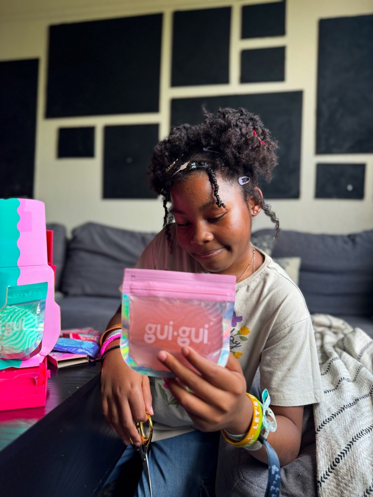 A young girl with curly hair is sitting on a couch, holding a colorful package labeled 'gui-gui.' She appears to be in a relaxed setting with other colorful packages nearby.