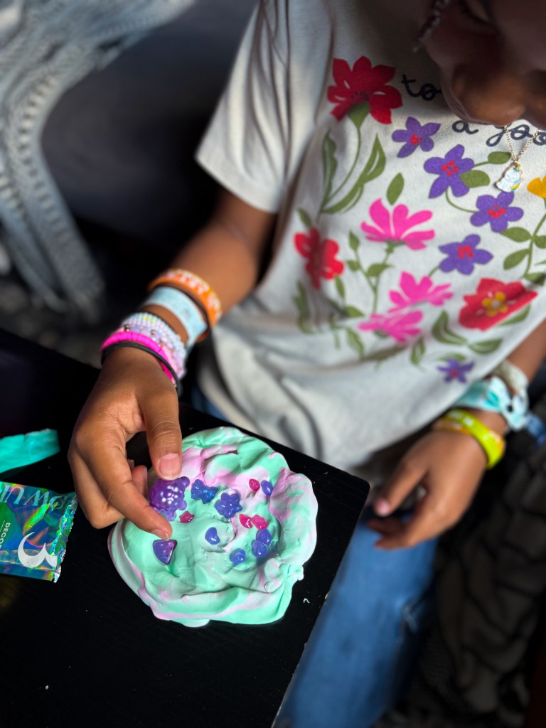 A child playing with colorful playdough, creating shapes and designs on a black table. The child is wearing a floral t-shirt and multiple wristbands.