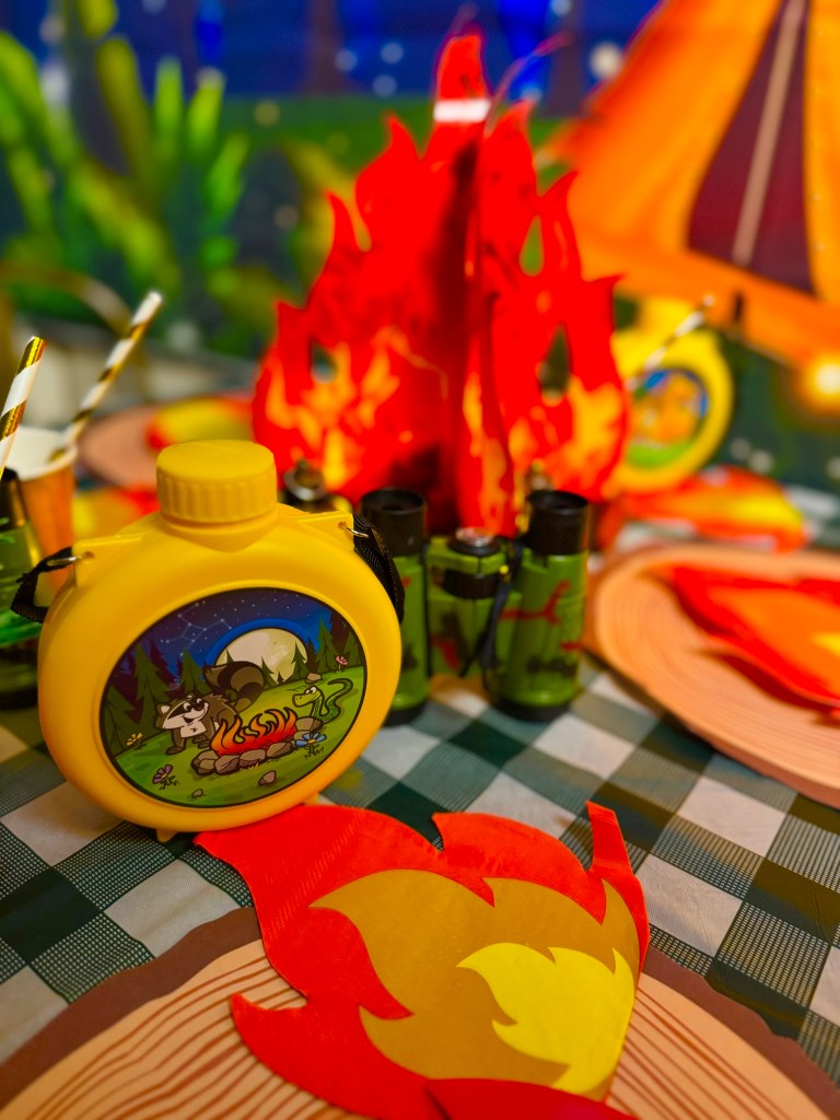 Camping party decorations with toy canteens, binoculars, paper pine trees, and cozy lantern lights.