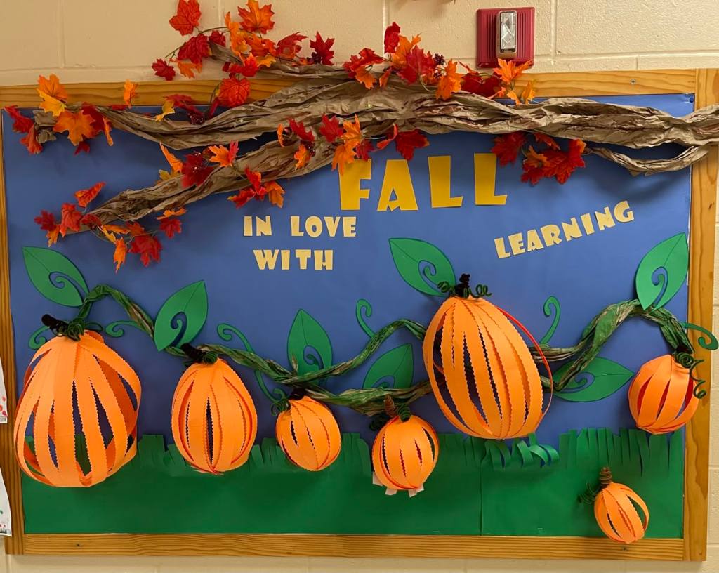 A decorative classroom bulletin board featuring orange paper pumpkins, green leaves, and a colorful autumn theme with the phrase 'FALL IN LOVE WITH LEARNING' in bold letters.