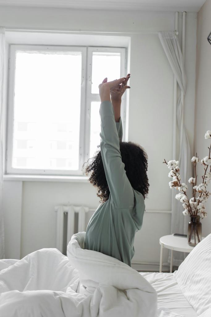 A person stretching in bed while surrounded by white blankets and a cozy room with a window.